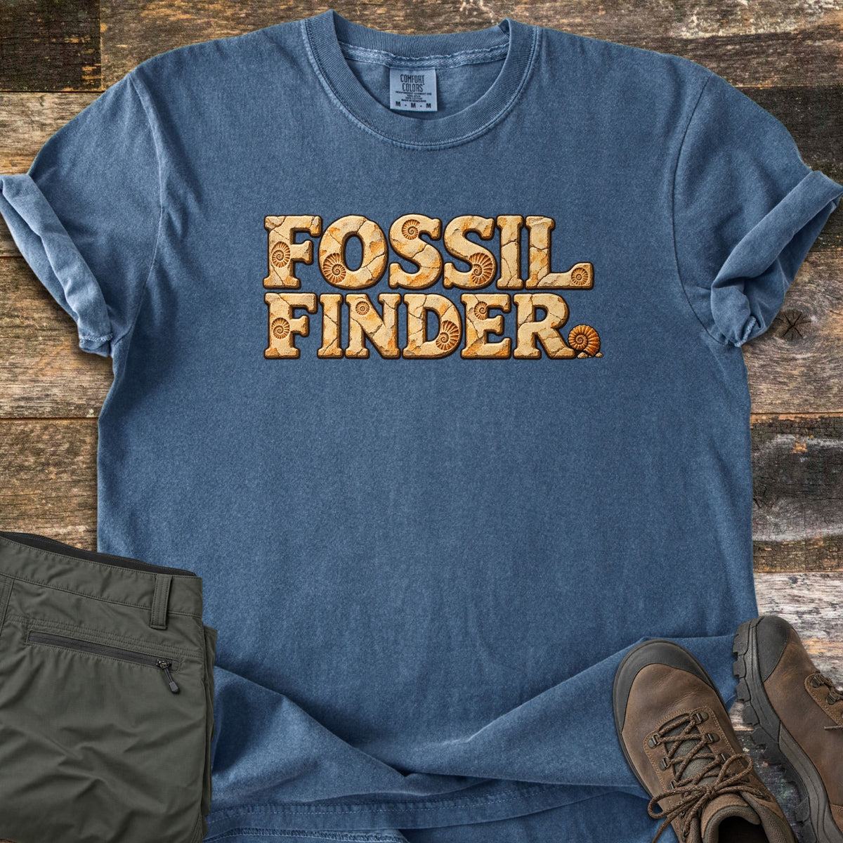 Fossil Finder Comfort Colors T-shirt in Denim, laid flat. The words Fossil Finder are printed in cracked, sandstone-textured letters with small ammonite fossils, ending with an ammonite shell. Green cargo pants and brown hiking boots sit beside it on weathered wood.