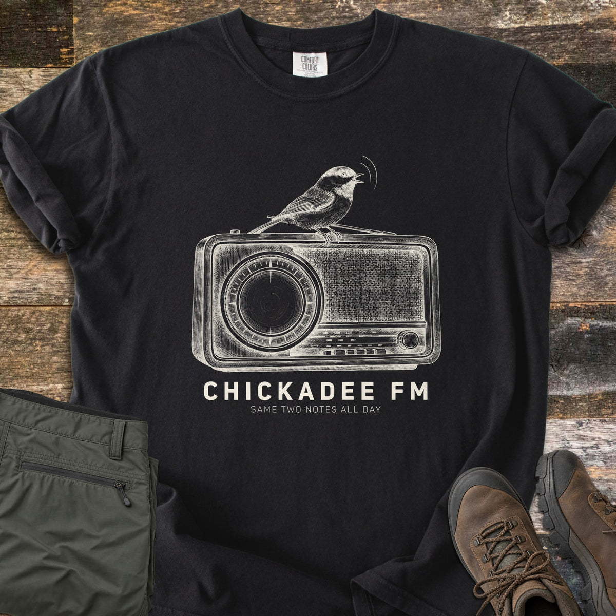 Chickadee FM Comfort Colors T-shirt in Black, laid flat. A sketched chickadee singing atop a vintage radio, above the words CHICKADEE FM and Same Two Notes All Day in cream. Sleeves rolled, on weathered wood with folded green hiking pants and brown hiking boots.