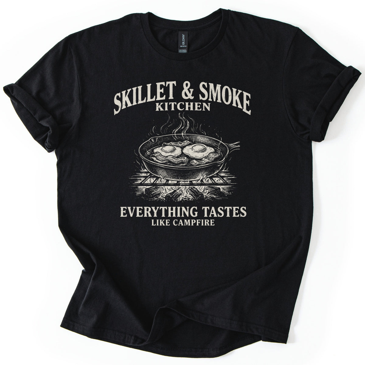 Skillet & Smoke Kitchen Comfort Colors T-shirt in Black, laid flat. A cream print shows a cast iron skillet of fried eggs and bacon steaming on a grate over a campfire, under the words Skillet & Smoke Kitchen and above Everything Tastes Like Campfire. Sleeves cuffed, on white.