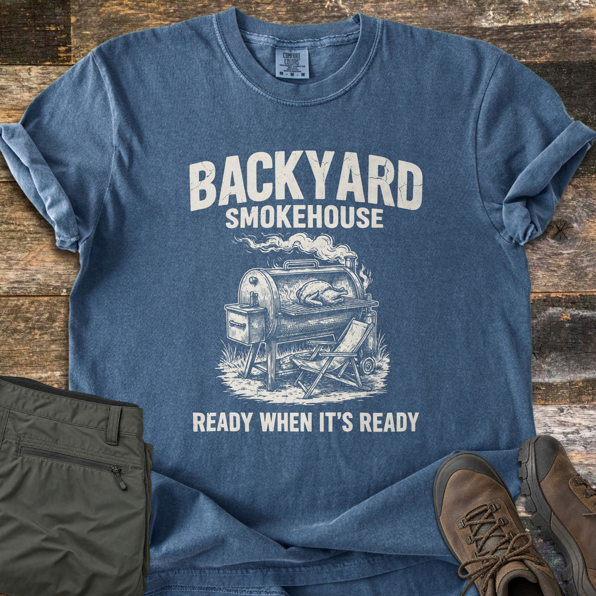 Backyard Smokehouse Comfort Colors T-shirt in Denim, laid flat. Distressed text reads Backyard Smokehouse above a sketch of a barrel smoker with a bird on the grate and a folding chair, then Ready When It's Ready. Laid on wood planks with olive pants and brown hiking boots.