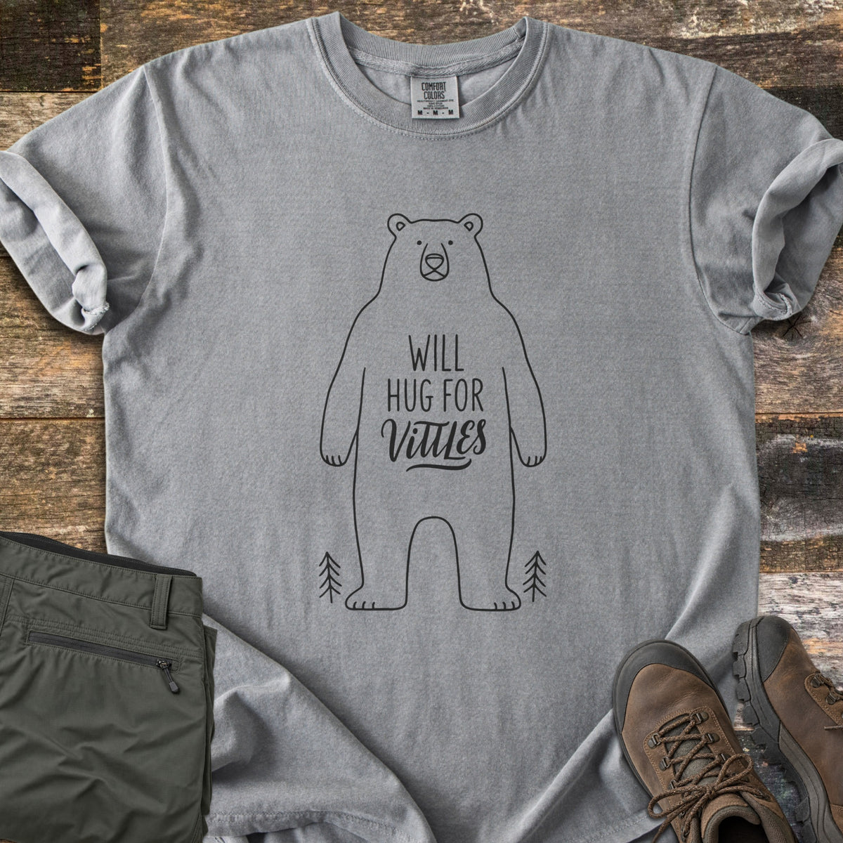 Will Hug For Vittles Comfort Colors T-shirt in Grey, laid flat. A black outline of a standing bear has the words Will Hug For Vittles on its belly, with two small pine trees by its feet. Folded olive hiking pants and brown hiking boots sit beside it on weathered wood.