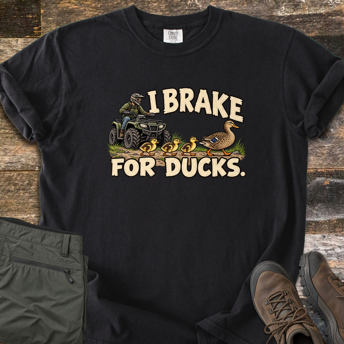 I Brake for Ducks Comfort Colors T-shirt in Black, laid flat. A rider in a helmet on a green ATV waits as a mother duck leads her ducklings across a dirt path, under the words I Brake For Ducks. Olive pants and brown hiking boots lie beside it on weathered wood planks.