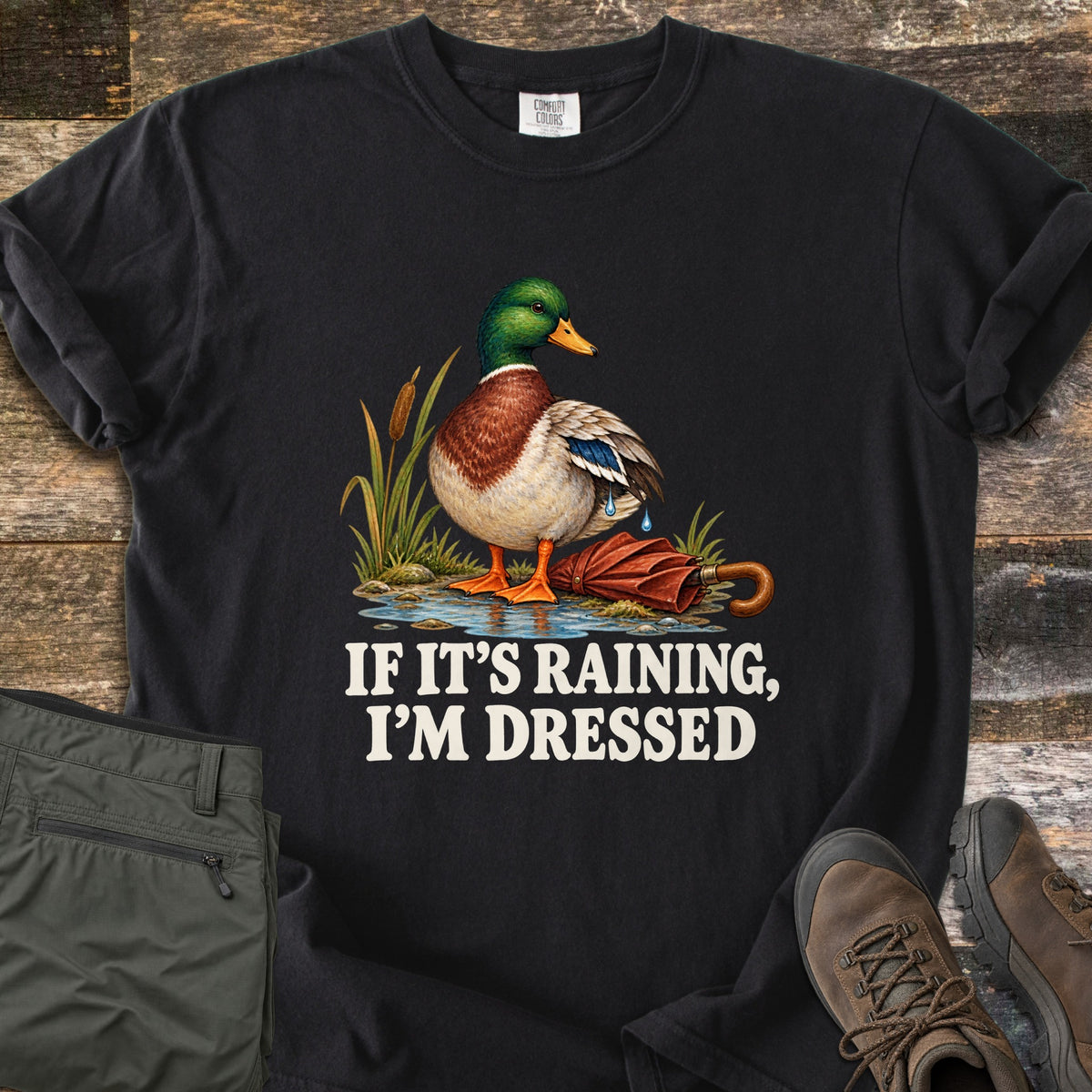 If It's Raining, I'm Dressed Comfort Colors T-shirt in Black, laid flat. A mallard duck stands in a puddle by cattails, water dripping off its wing, next to a closed red umbrella, above the words If It's Raining, I'm Dressed in white. Olive pants and brown hiking boots lie on weathered wood.