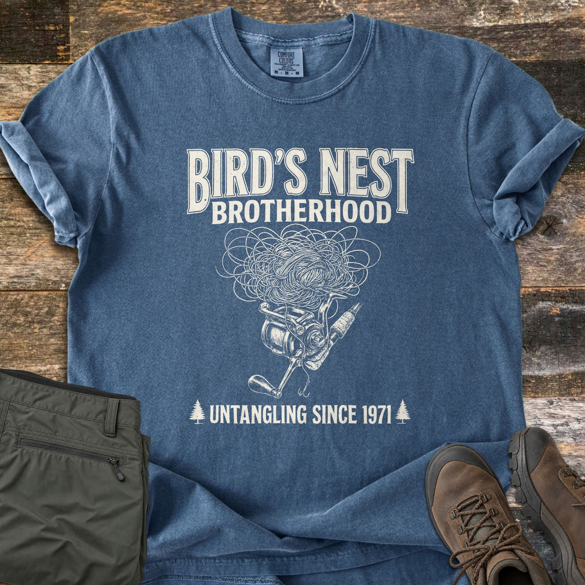 Birds Nest Brotherhood Comfort Colors T-shirt in Denim, laid flat. Cream print reads Bird's Nest Brotherhood above a fishing reel and rod with a tangled ball of line, then Untangling Since 1971 between two pine trees. Shown on wood with olive pants and brown hiking boots.
