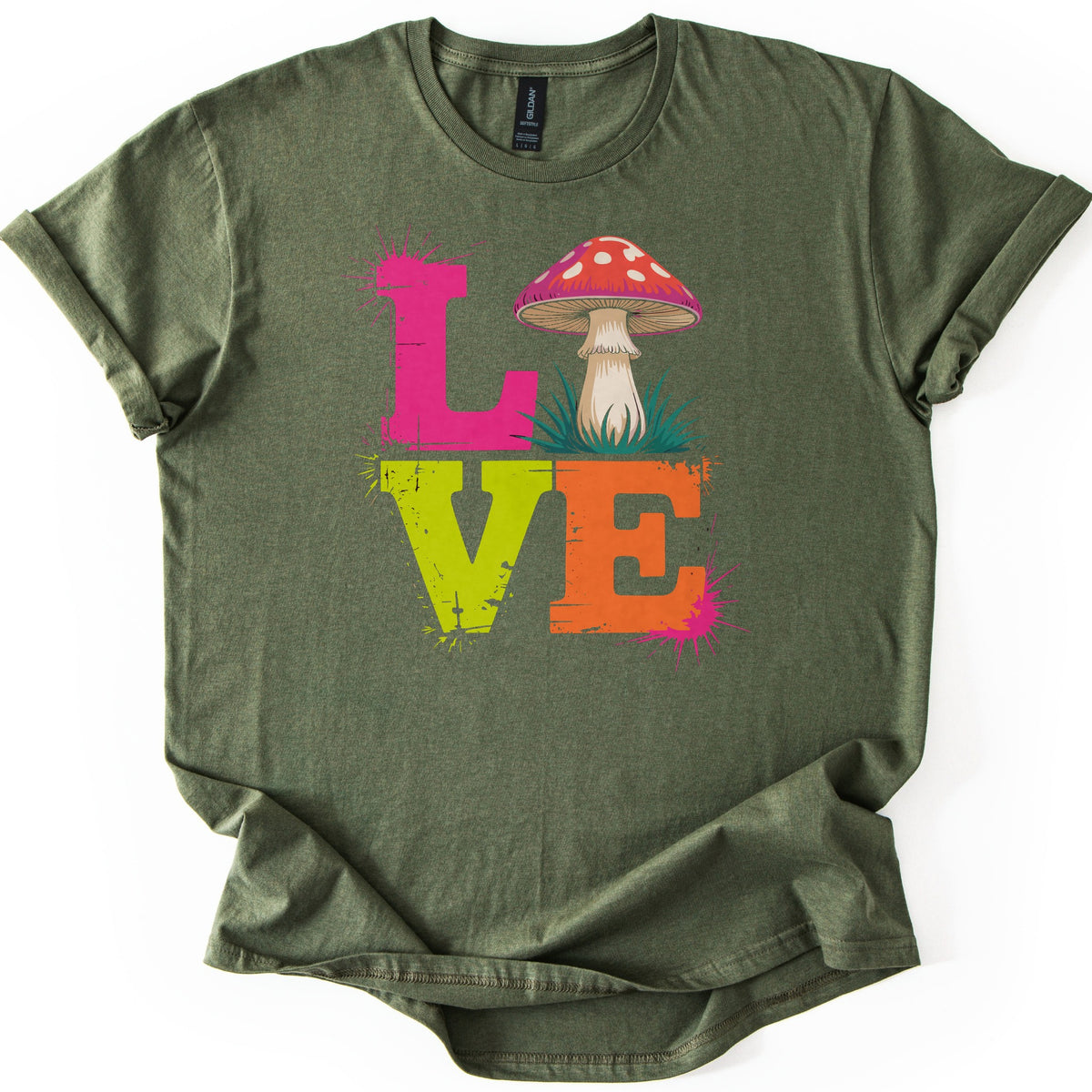 Mushroom Love Comfort Colors T-shirt in Military Green, laid flat. The word LOVE is printed in distressed block letters, a pink L, a red spotted mushroom in grass replacing the O, a lime green V and an orange E, with pink splatters. The shirt lies on white with rolled sleeves.