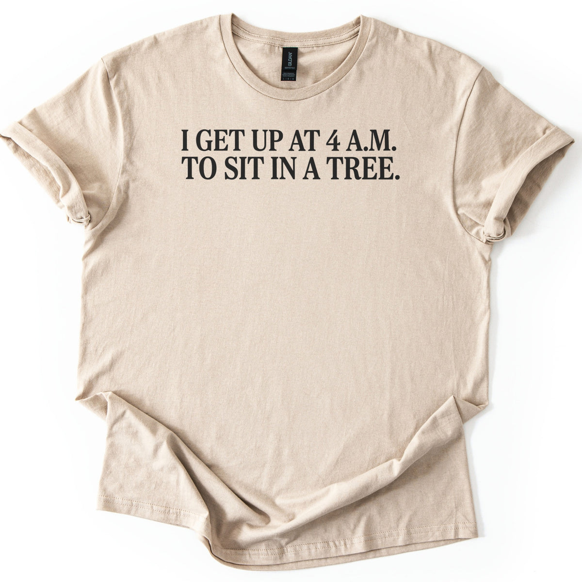 Sit In A Tree Comfort Colors T-shirt in Sand, laid flat. Black serif capital letters across the chest read I GET UP AT 4 A.M. TO SIT IN A TREE. on two lines. The shirt lies on a white surface with its sleeves rolled and the hem loosely bunched.