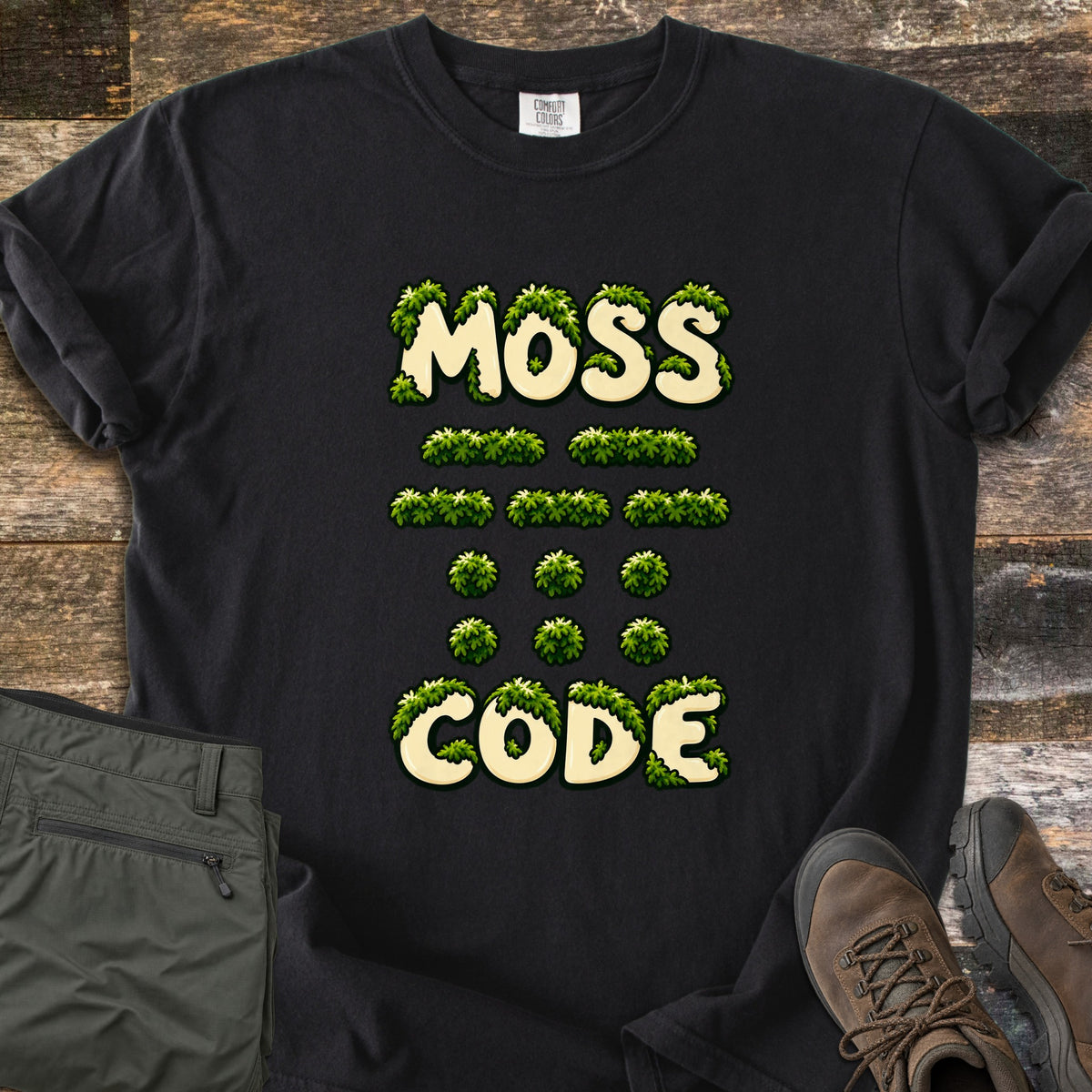 Moss Code Comfort Colors T-shirt in Black, laid flat. Moss-covered cream bubble letters spell MOSS, then moss-tuft dashes and dots spelling MOSS in Morse code, then CODE. It lies on weathered wood beside folded olive hiking pants and a brown hiking boot.