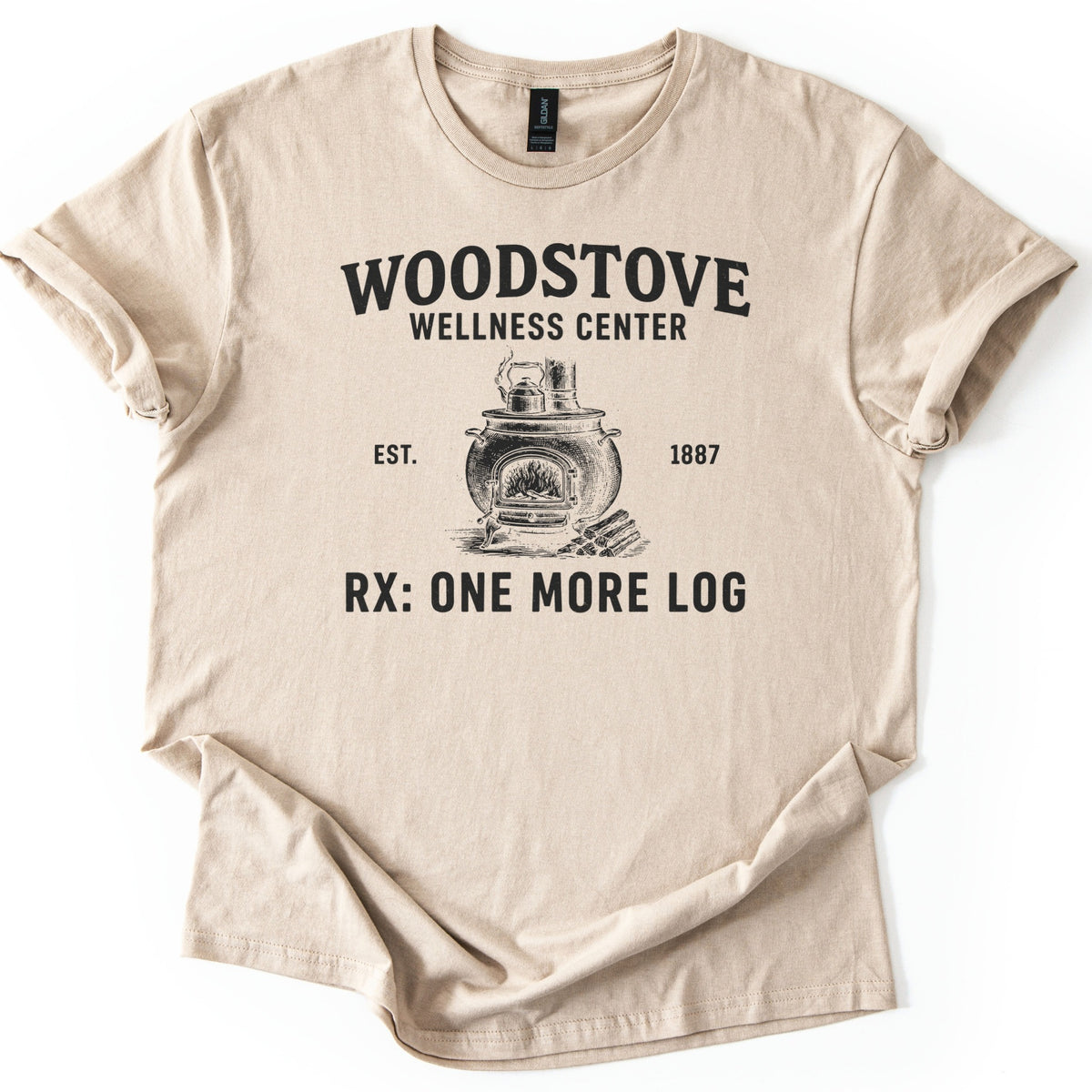 Woodstove Wellness Center Comfort Colors T-shirt in Sand, laid flat. A black sketch of a round woodstove with a kettle on top, flames in its open door and stacked logs beside it, under Woodstove Wellness Center, Est. 1887, and Rx: One More Log. Sleeves rolled, on white.