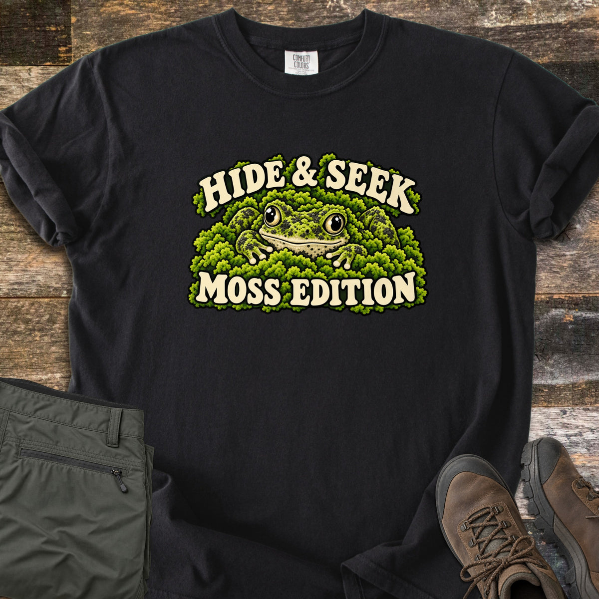 Hide & Seek Moss Edition Comfort Colors T-shirt in Black, laid flat. A wide-eyed frog peeks out from a mound of green moss between the words Hide & Seek and Moss Edition. Olive cargo pants and brown hiking boots lie beside it on weathered wood planks.