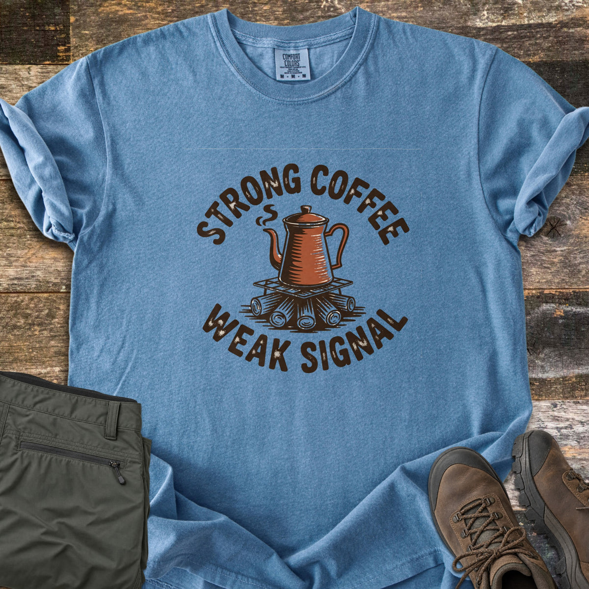 Strong Coffee Weak Signal Comfort Colors T-shirt in Blue Jean, laid flat. A steaming copper coffee pot sits on a grate over campfire logs, arched by the words Strong Coffee above and Weak Signal below. Folded olive hiking pants and brown hiking boots lie on weathered wood planks.