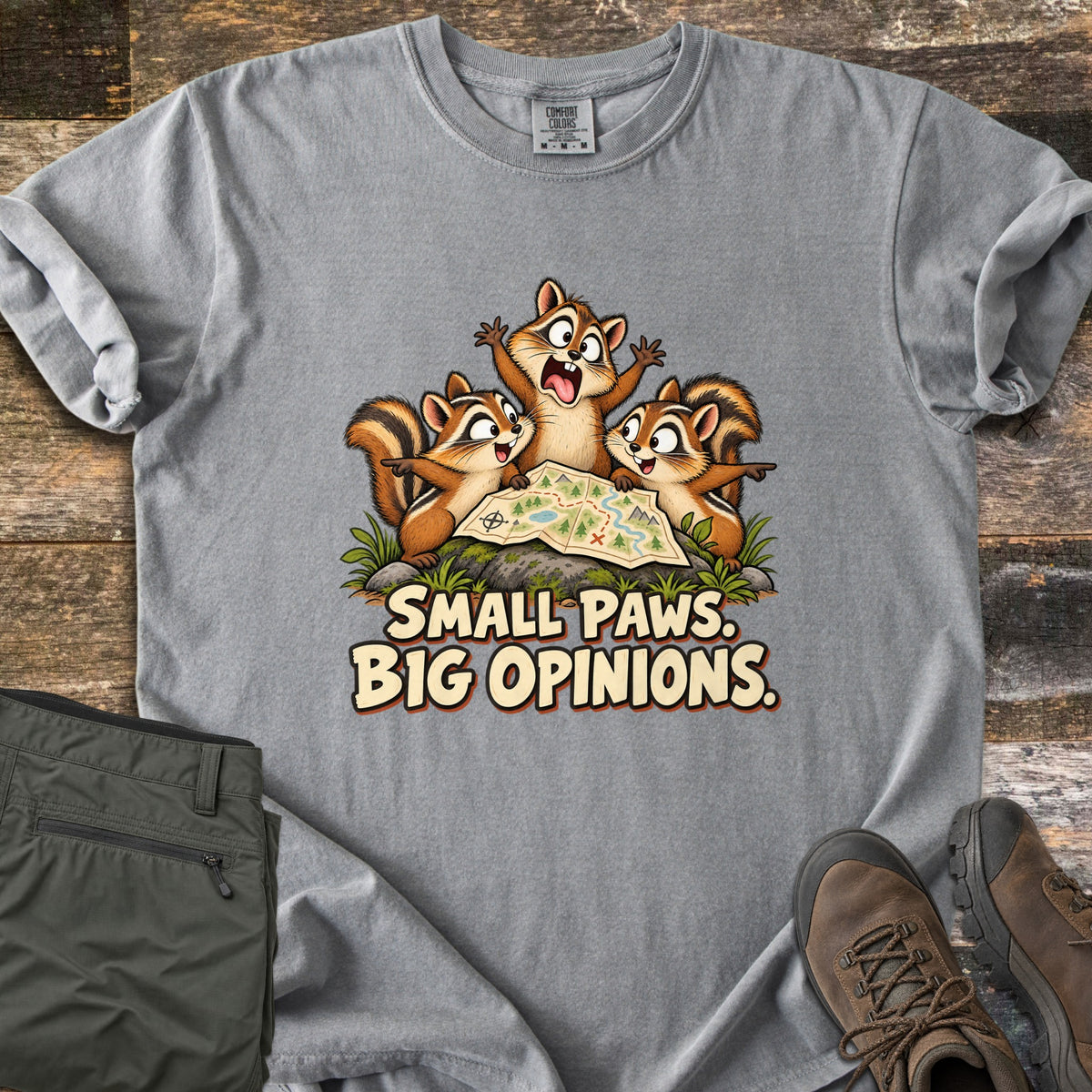 Small Paws. Big Opinions. Comfort Colors T-shirt in Grey, laid flat. Three cartoon chipmunks argue over a trail map on a mossy rock, one with arms thrown up, above the words SMALL PAWS. BIG OPINIONS. Olive hiking pants and brown hiking boots sit beside it on dark wood planks.
