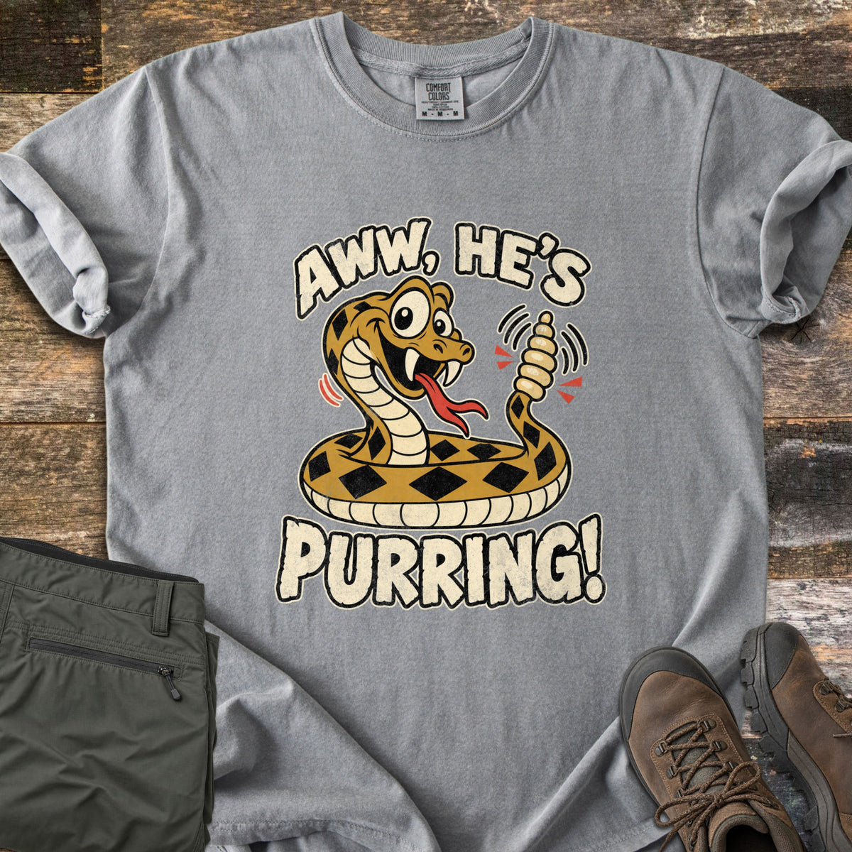 Aww He's Purring Comfort Colors T-shirt in Grey, laid flat. A cartoon rattlesnake with a red forked tongue and motion lines around its rattle sits between the words Aww, He's Purring! Folded olive pants and brown hiking boots rest beside it on dark wood planks.