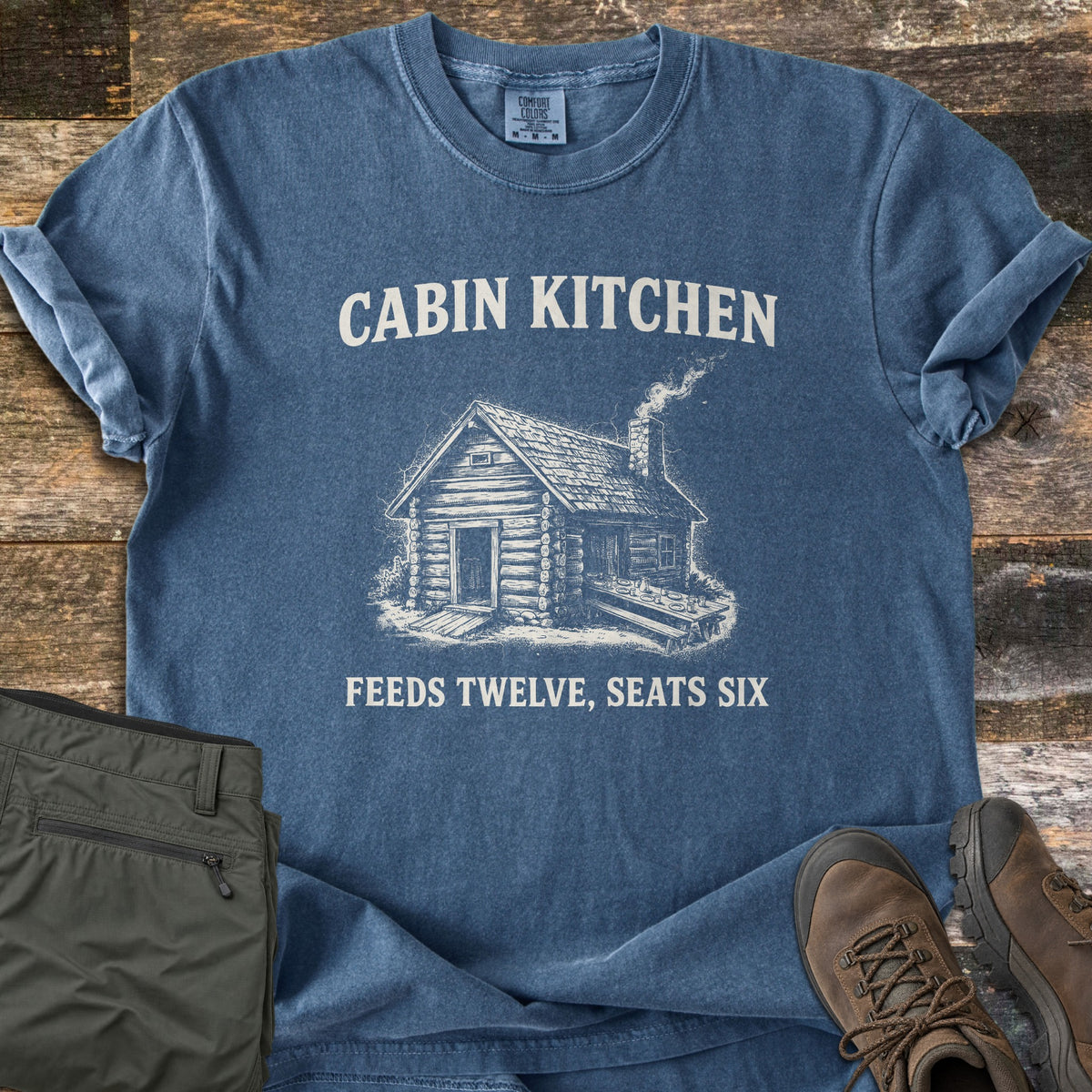 Cabin Kitchen Comfort Colors T-shirt in Denim, laid flat. A white sketch of a smoking log cabin with a long picnic table sits between the words Cabin Kitchen and Feeds Twelve, Seats Six. Olive hiking pants and brown hiking boots lie beside it on weathered wood planks.