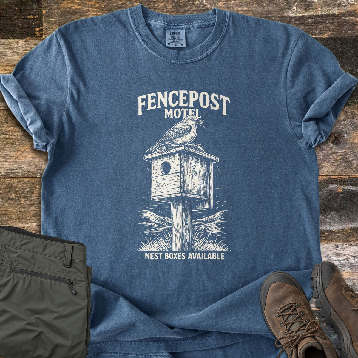 Fencepost Motel Comfort Colors T-shirt in Denim, laid flat. Cream print of a bird on a wooden birdhouse atop a post in grass, with the words Fencepost Motel above and Nest Boxes Available below. On a wood floor beside olive cargo pants and brown hiking boots.
