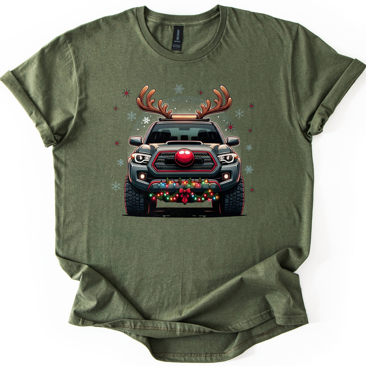 Santa Truck Comfort Colors T-shirt in Military Green, laid flat. A dark gray pickup truck seen head-on wears reindeer antlers and a red nose, with colored string lights and a red bow on its bumper, framed by snowflakes and red stars. No words. Sleeves cuffed, on white.