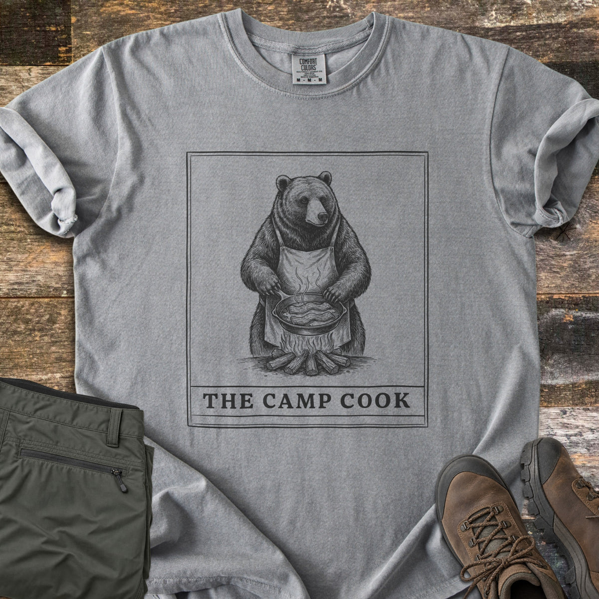 The Camp Cook Comfort Colors T-shirt in Grey, laid flat. A framed sketch of a bear in an apron frying bacon in a skillet over a campfire, above the words THE CAMP COOK. Sleeves are cuffed on weathered wood beside olive hiking pants and brown hiking boots.