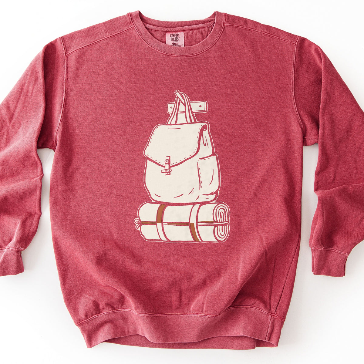 Backpack And Bedroll Comfort Colors crewneck sweatshirt in Crimson, laid flat. A cream canvas backpack with a toggle clasp hangs by its straps from a wall peg above a rolled cream bedroll tied with brown straps. No words are printed. Shown on a white background.