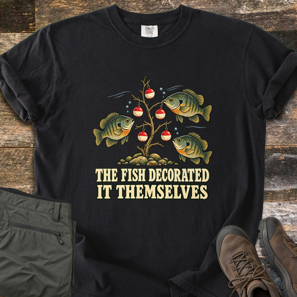 The Fish Decorated It Themselves Comfort Colors T-shirt in Black, laid flat. Three bluegill fish hang red and white fishing bobbers like ornaments on a bare underwater branch above the words The Fish Decorated It Themselves. Olive pants and brown hiking boots sit on weathered wood.