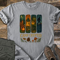 Camp Cryptid Big Foot Comfort Colors T-shirt in Grey, laid flat. Four painted panels show Bigfoot at a campfire, a lantern, a backpack marked GONE with binoculars, and Bigfoot under stars, above CAMP CRYPTID, a moth, pinecone and mushrooms. Olive pants and hiking boots lie on wood.