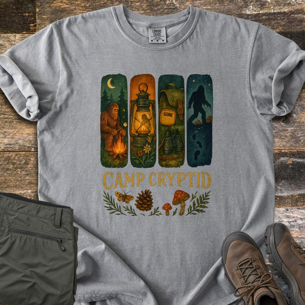 Camp Cryptid Big Foot Comfort Colors T-shirt in Grey, laid flat. Four painted panels show Bigfoot at a campfire, a lantern, a backpack marked GONE with binoculars, and Bigfoot under stars, above CAMP CRYPTID, a moth, pinecone and mushrooms. Olive pants and hiking boots lie on wood.