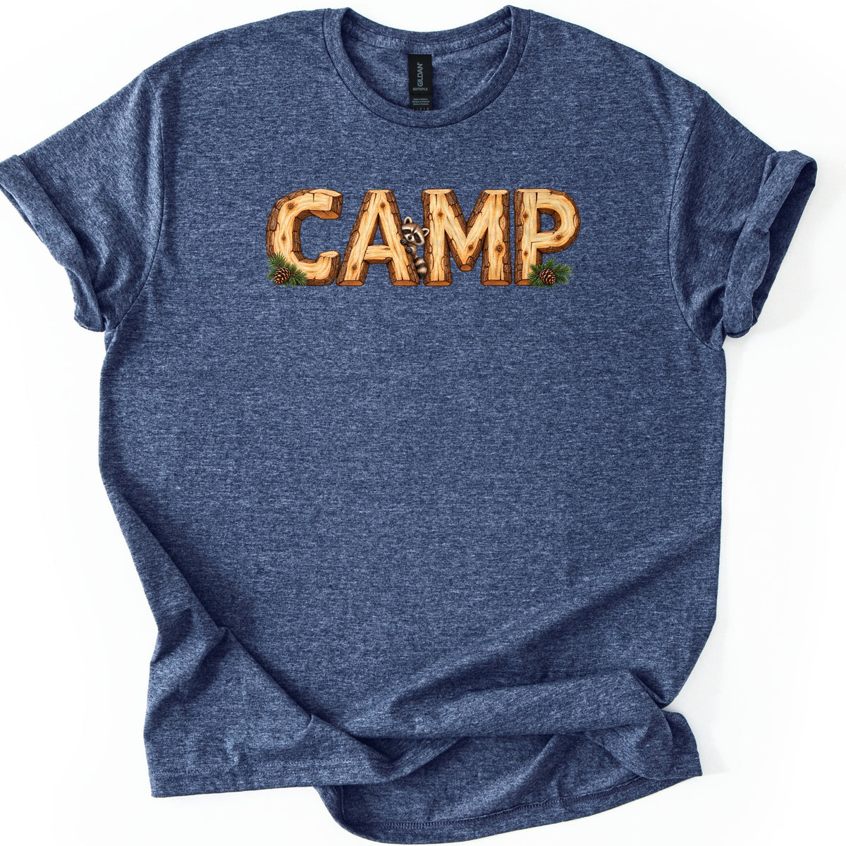 Camp Comfort Colors T-shirt in Heather Navy, laid flat. The word CAMP is printed in log letters with bark edges, a raccoon peeking out between the A and M, and pine sprigs with pinecones at both ends. The heathered shirt lies on a white background with cuffed sleeves.
