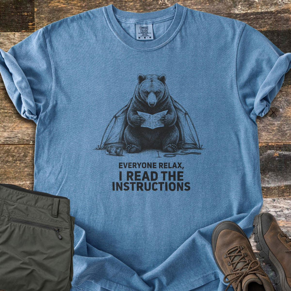Everyone Relax I Read The Instructions Comfort Colors T-shirt in Blue Jean, laid flat. A black sketch of a bear reading instructions in front of a tent, with a tipped lantern and rope, above the words Everyone relax, I read the instructions. Olive cargo pants and brown hiking boots sit on dark wood.