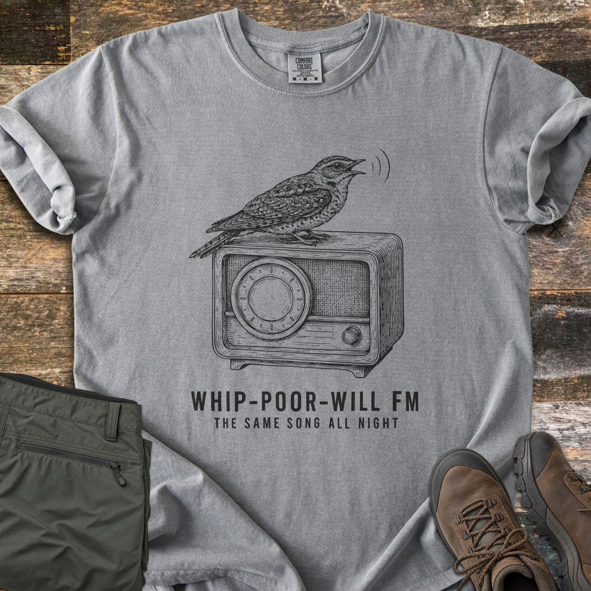 Whip-Poor-Will FM Comfort Colors T-shirt in Grey, laid flat. A sketched whip-poor-will sings atop a vintage radio above the words Whip-Poor-Will FM, The Same Song All Night. It lies with rolled sleeves on weathered wood beside folded olive pants and brown hiking boots.