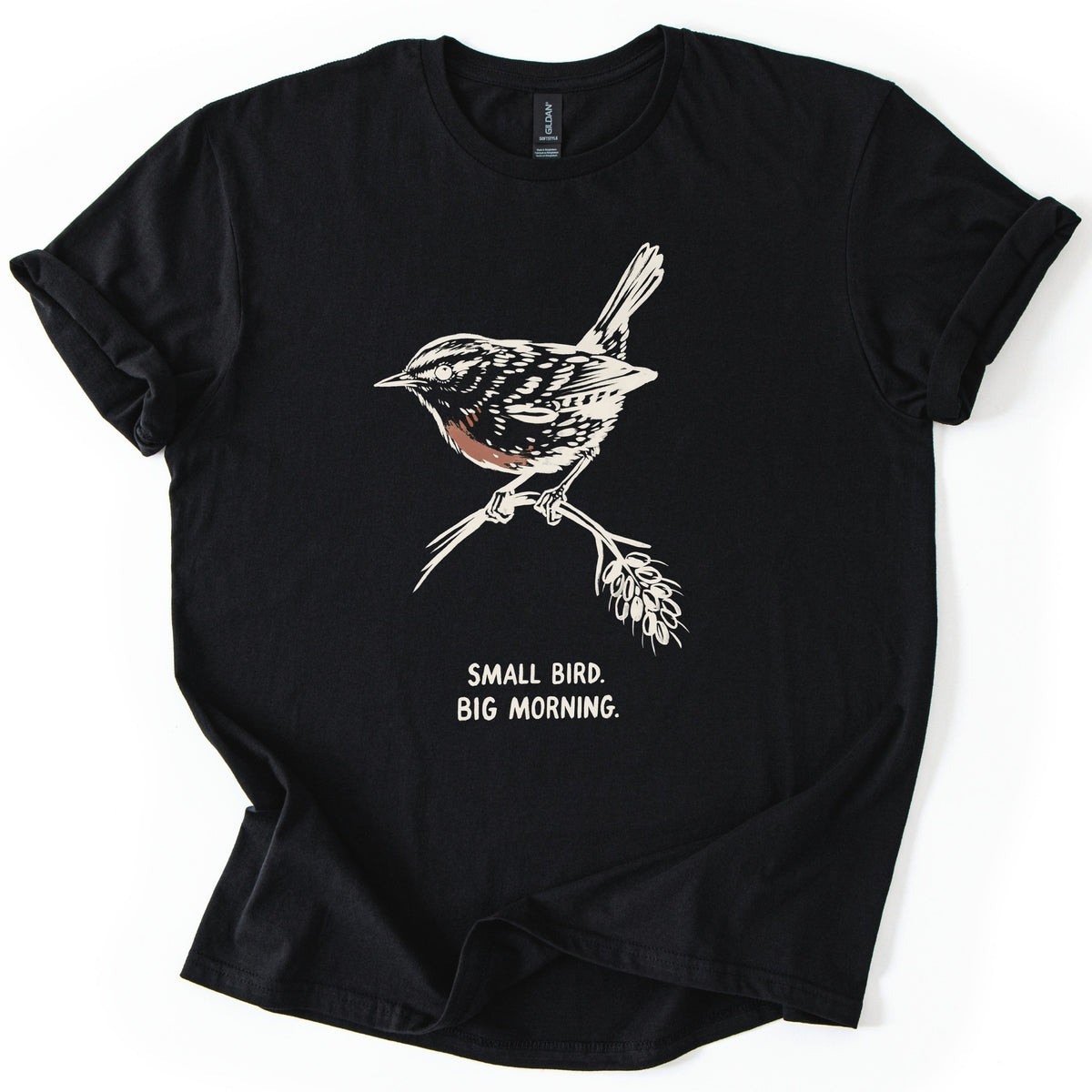 Small Bird Big Morning Comfort Colors T-shirt in Black, laid flat. A cream sketch of a wren with a rust-colored breast perches on a twig with a seed head, above the words Small Bird. Big Morning. The sleeves are rolled up, on a white background.