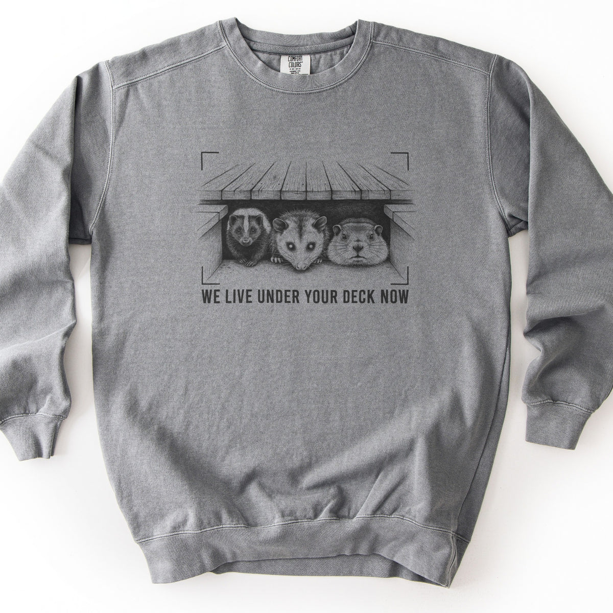 We Live Under Your Deck Now Comfort Colors crewneck sweatshirt in Grey, laid flat. A black sketch of a skunk, opossum and groundhog looking out from under a wooden deck, framed by viewfinder-style corner brackets, sits above the words We Live Under Your Deck Now. Plain white background.