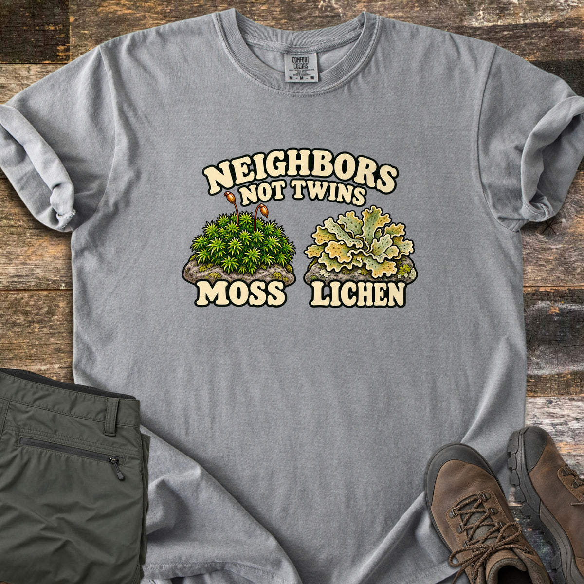 Neighbors Not Twins Comfort Colors T-shirt in Grey, laid flat. Arched text Neighbors Not Twins over two illustrated rock clumps: green moss with brown stalks labeled Moss, and pale green leafy lichen labeled Lichen. Olive hiking pants and brown boots sit beside it on weathered wood.