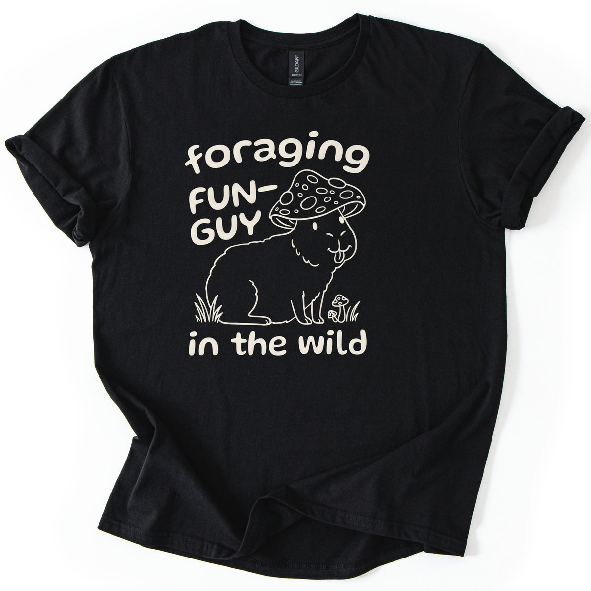 Foraging Fun-Guy Capybara Comfort Colors T-shirt in Black, laid flat. A cream line drawing of a winking capybara with its tongue out, wearing a spotted mushroom cap and sitting in grass by tiny mushrooms, with the words foraging FUN-GUY in the wild. Sleeves rolled, on white.