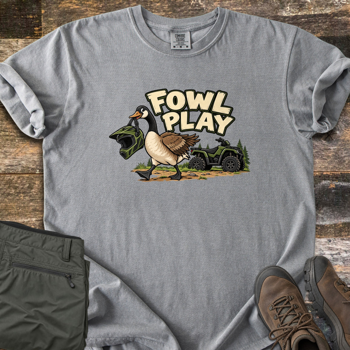 Fowl Play Comfort Colors T-shirt in Grey, laid flat. A cartoon Canada goose carries a green off-road helmet in its beak past a green ATV and pine trees, under the words Fowl Play. Sleeves rolled, on weathered wood with olive pants and brown hiking boots.