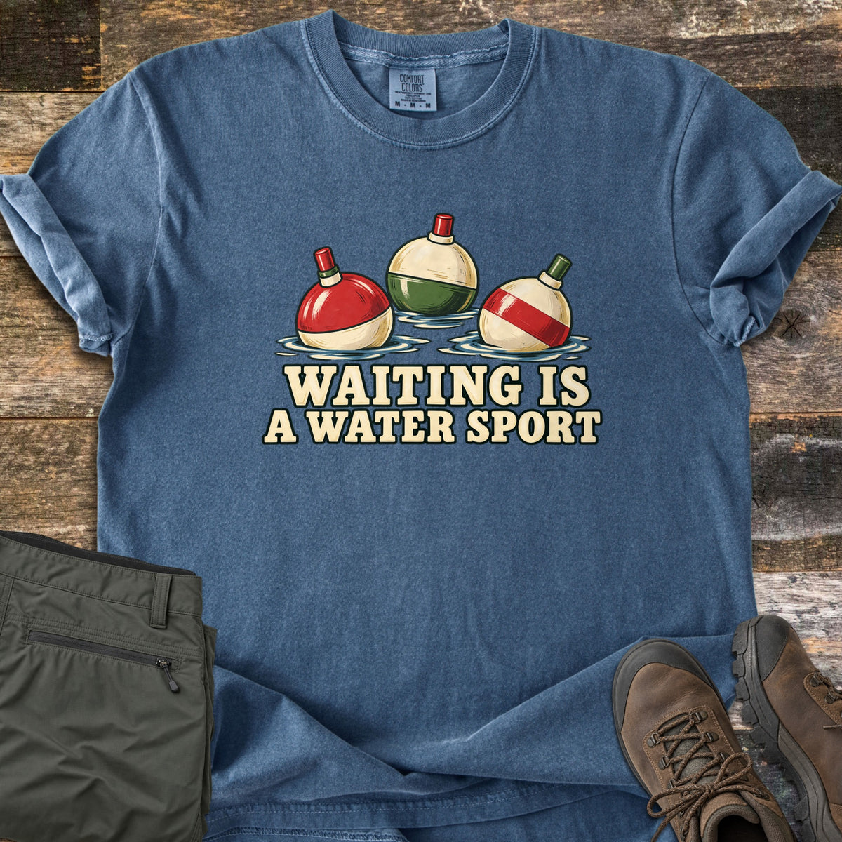 Waiting Is a Water Sport Comfort Colors T-shirt in Denim, laid flat. Three red, cream, and green fishing bobbers float in rippled water above the words Waiting Is a Water Sport. Olive pants and brown hiking boots sit beside it on weathered wood planks.