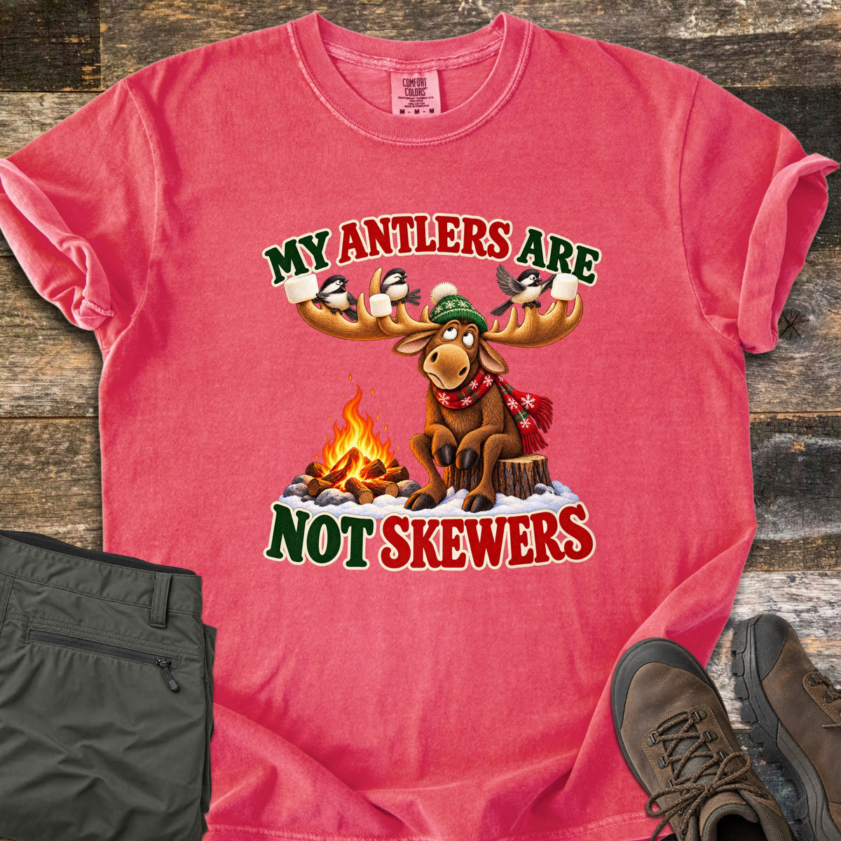 My Antlers Are Not Skewers Comfort Colors T-shirt in Watermelon, laid flat. A cartoon moose in a green knit hat and red scarf sits on a stump by a campfire as chickadees on its antlers hold marshmallows, with the words My Antlers Are Not Skewers. Olive pants and brown boots lie on weathered wood.