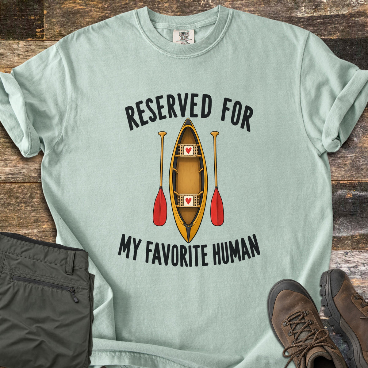Reserved For My Favorite Human Comfort Colors T-shirt in Bay, laid flat. A yellow canoe seen from above with a red heart on each seat sits between two red-bladed paddles, under the words Reserved For and above My Favorite Human. Olive pants and brown hiking boots lie beside it on wood planks.