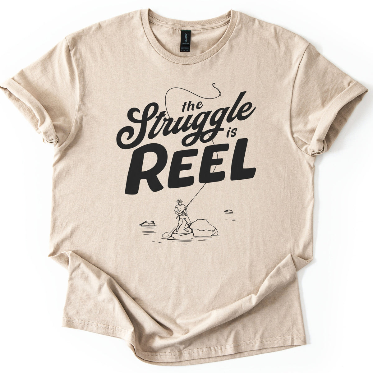 Struggle Is Reel Angler Comfort Colors T-shirt in Sand, laid flat. Black text reads the Struggle is REEL, with a fishing line arcing over the words to a hook. Below, a line drawing of an angler in a hat casts from a rock in the water. Sleeves cuffed, on a white background.