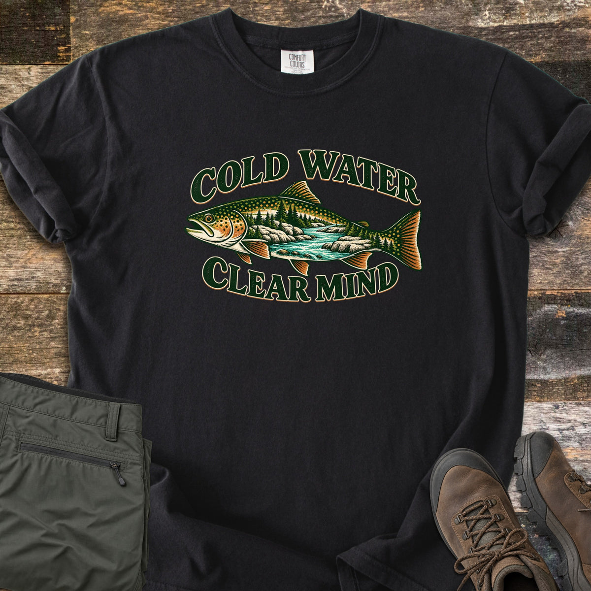 Cold Water Clear Mind Comfort Colors T-shirt in Black, laid flat. A spotted trout with a forest and turquoise stream inside its body sits between the green outlined words Cold Water and Clear Mind. Folded olive hiking pants and brown hiking boots lie beside it on old wood planks.