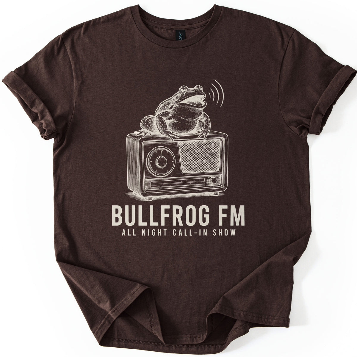 Bullfrog FM Comfort Colors T-shirt in Dark Chocolate, laid flat. A cream ink sketch of a croaking bullfrog on a vintage radio, sound waves at its mouth, above the words BULLFROG FM and ALL NIGHT CALL-IN SHOW. Sleeves are cuffed on a white background.