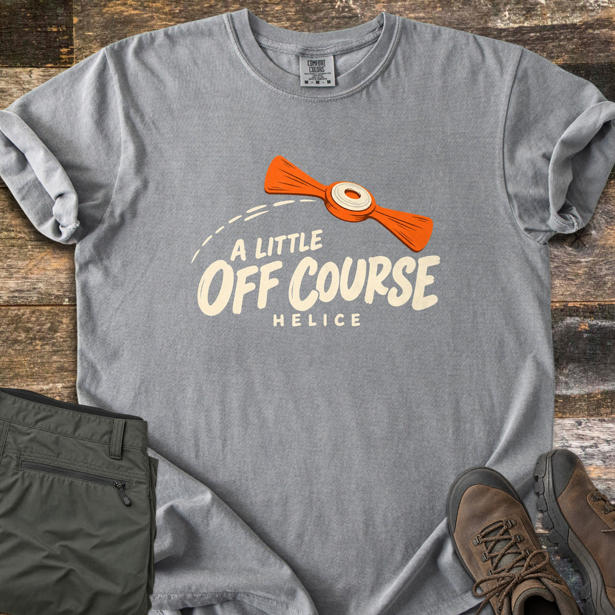 A Little Off Course Comfort Colors T-shirt in Grey, laid flat. An orange helice target with a white center flies along a dashed arc above cream text reading A Little Off Course and Helice. Folded olive hiking pants and brown hiking boots sit beside it on weathered wood.