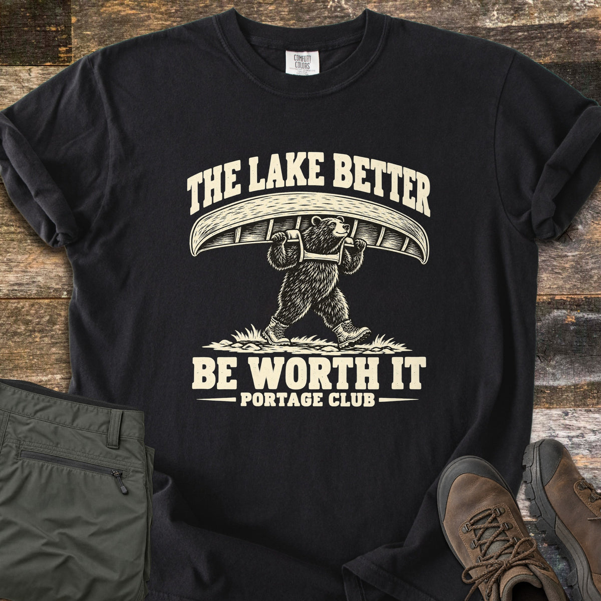 The Lake Better Be Worth It Comfort Colors T-shirt in Black, laid flat. A bear in hiking boots carries a canoe overhead between the words The Lake Better Be Worth It, with Portage Club below, printed in cream. Shown on weathered wood with folded green pants and brown hiking boots.