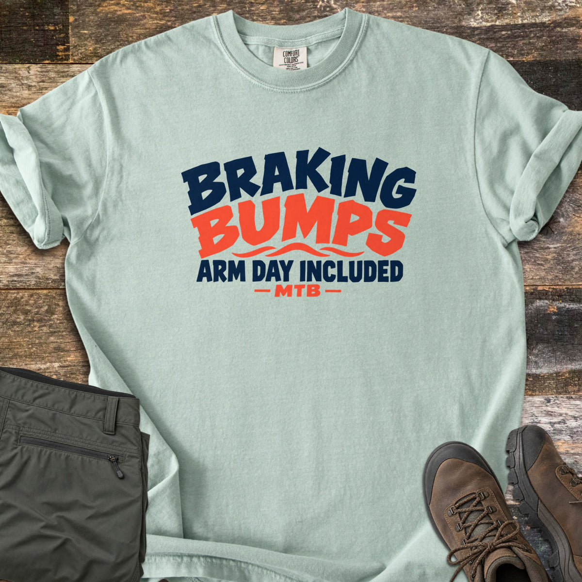 Braking Bumps Arm Day Included Comfort Colors T-shirt in Bay, laid flat. Bold navy and orange text reads Braking Bumps, Arm Day Included, MTB, with wavy orange lines under Bumps. It rests on a wood floor beside olive cargo pants and brown hiking boots.