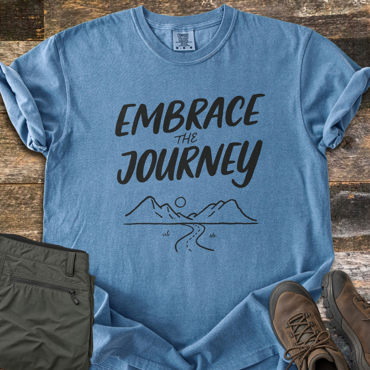 Embrace the Journey Comfort Colors T-shirt in Blue Jean, laid flat. Black brush lettering reads Embrace the Journey above a line drawing of mountains, a sun, and a winding road. Sleeves cuffed on weathered wood beside folded green pants and brown hiking boots.