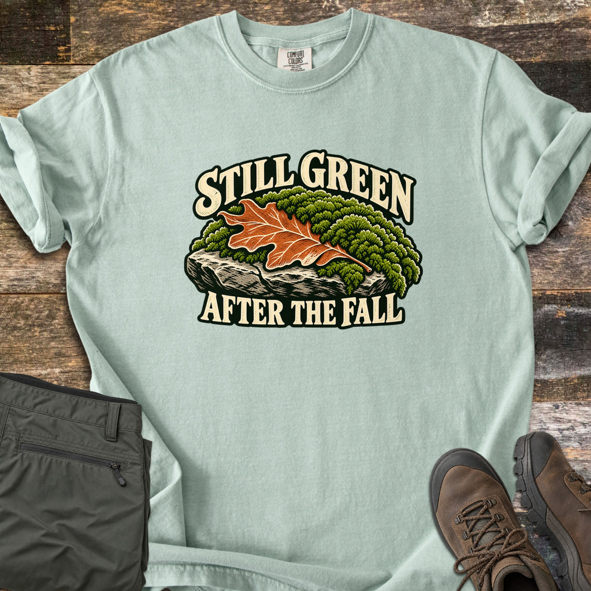 Still Green After the Fall Comfort Colors T-shirt in Bay, laid flat. An orange oak leaf sits on green moss atop a gray rock, with Still Green above and After the Fall below in cream letters. Folded olive hiking pants and brown hiking boots lie nearby on weathered wood planks.