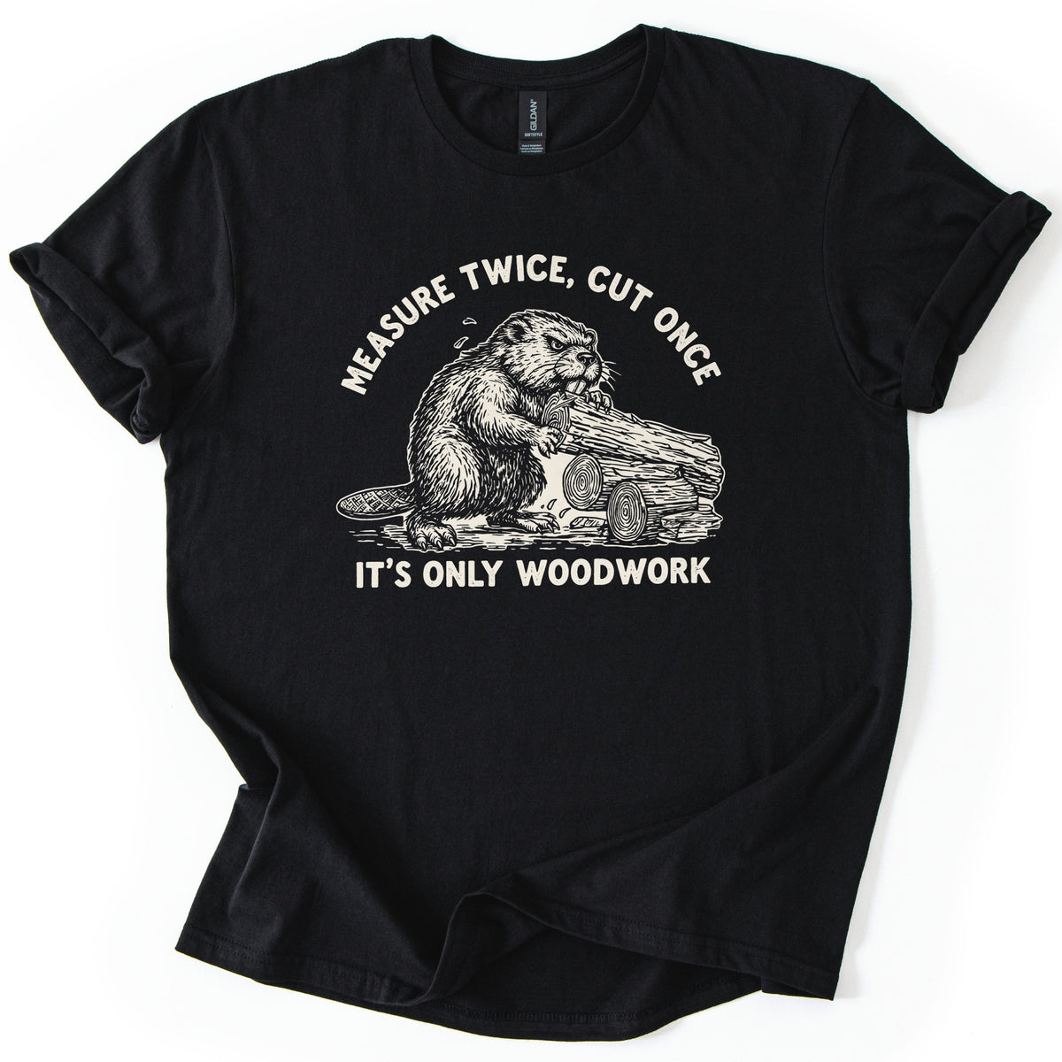 Measure Twice Beaver Comfort Colors T-shirt in Black, laid flat. A cream sketch of a beaver gnawing a log beside cut logs, under the arched words Measure Twice, Cut Once and above It's Only Woodwork. The shirt has rolled sleeves on a white background.