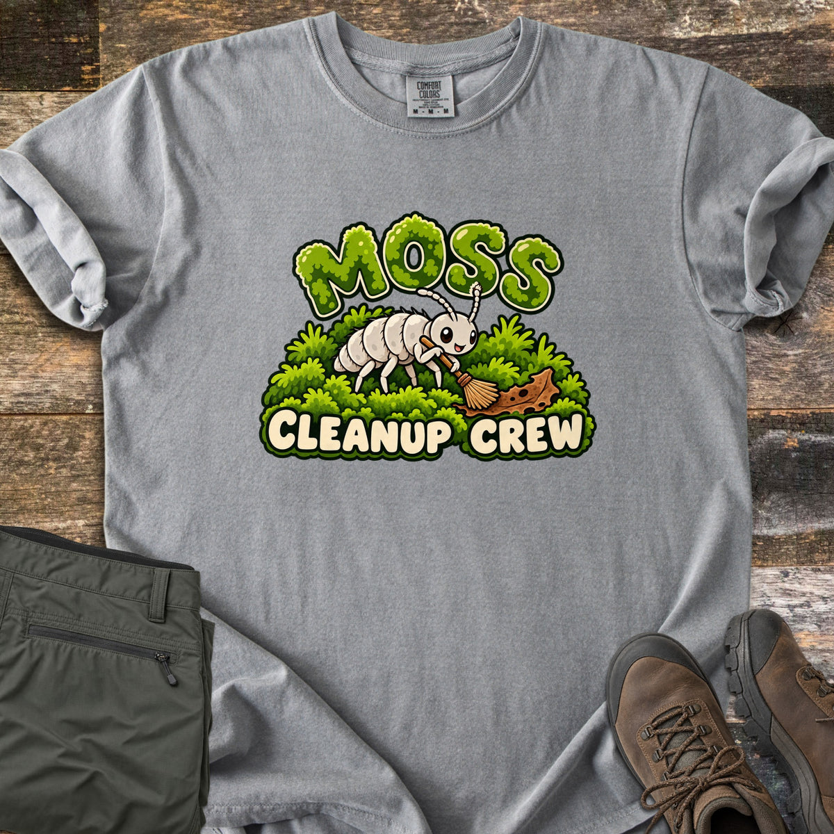 Moss Cleanup Crew Comfort Colors T-shirt in Grey, laid flat. A smiling white cartoon bug sweeps a brown leaf with a broom atop green moss, below the word MOSS in mossy letters and above CLEANUP CREW. It lies on weathered wood with folded olive pants and brown hiking boots.