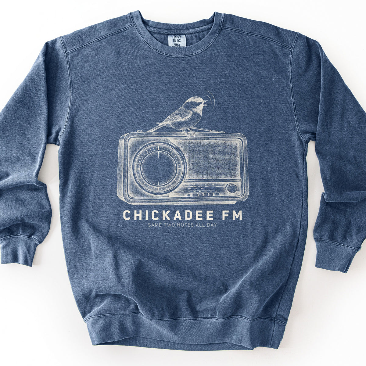 Chickadee FM Comfort Colors crewneck sweatshirt in Denim, laid flat. A vintage-style cream sketch of a chickadee singing atop an old radio, above the words CHICKADEE FM and SAME TWO NOTES ALL DAY. The sweatshirt lies on a plain white background.