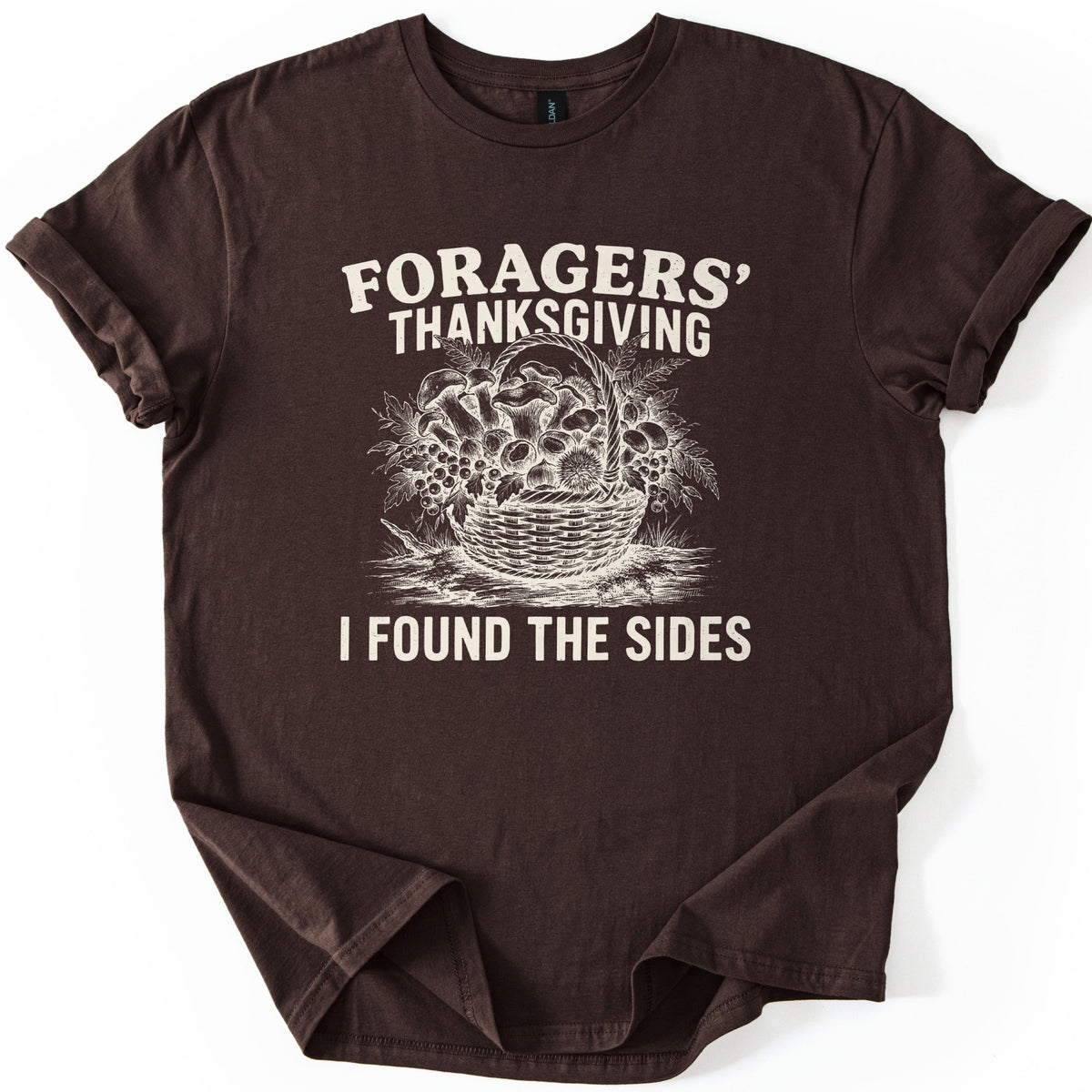 Foragers Thanksgiving Comfort Colors T-shirt in Dark Chocolate, laid flat. Cream sketch of a wicker basket of mushrooms, berries, ferns and a chestnut burr, between the words Foragers' Thanksgiving and I Found the Sides. Sleeves cuffed, on a white background.
