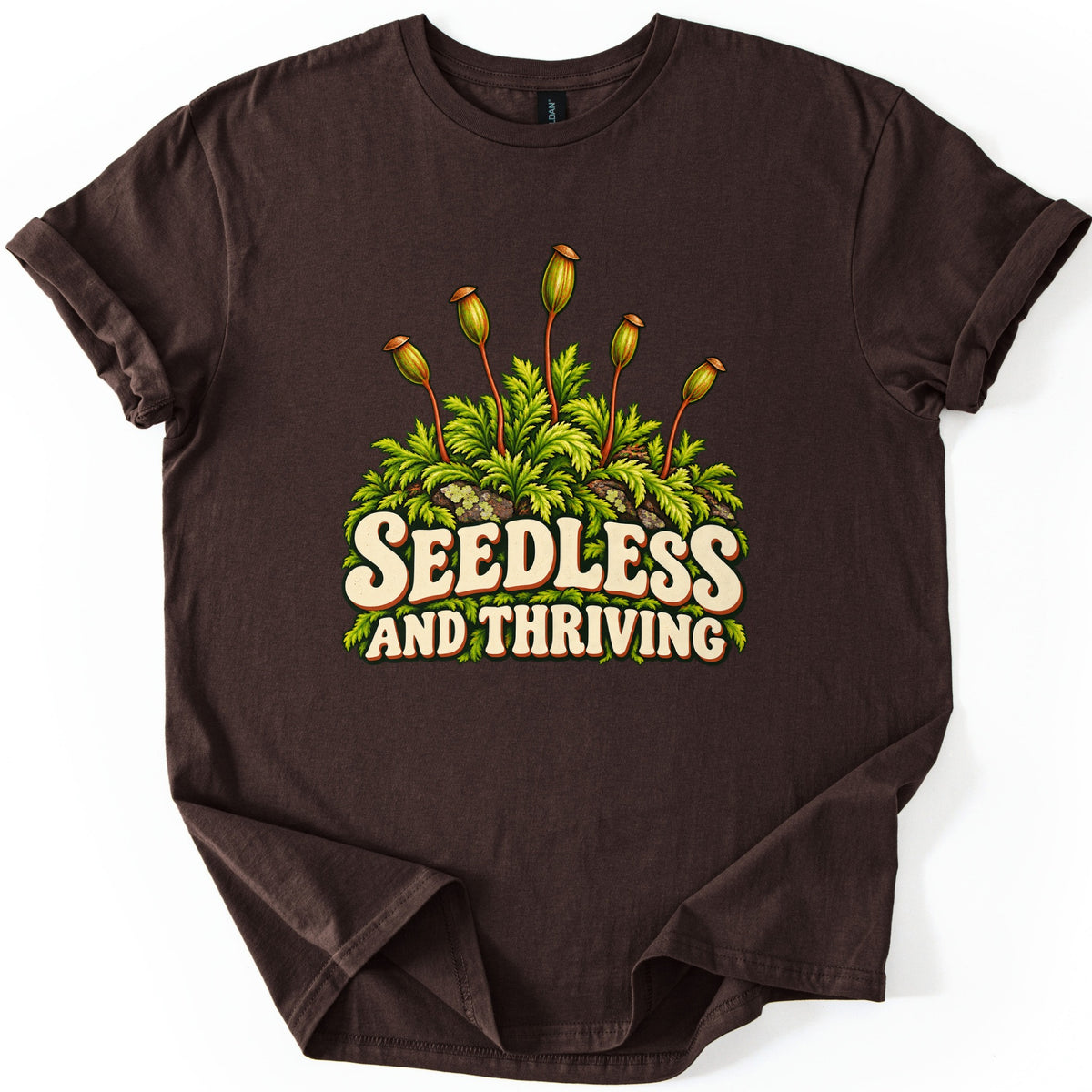 Seedless and Thriving Comfort Colors T-shirt in Dark Chocolate, laid flat. Green moss with tall reddish stalks topped by spore capsules grows over lichen-dotted rocks above the words SEEDLESS AND THRIVING in cream retro letters. The shirt lies on white with sleeves rolled.