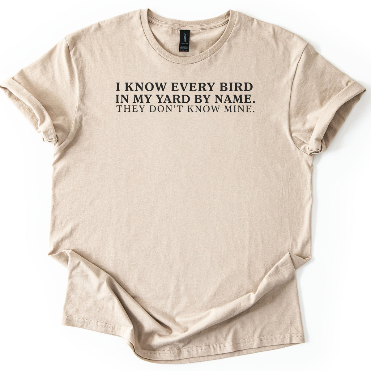 I Know Every Bird in My Yard by Name Comfort Colors T-shirt in Sand, laid flat. Black serif text across the chest reads I KNOW EVERY BIRD IN MY YARD BY NAME. THEY DON'T KNOW MINE. The sleeves are cuffed and the shirt lies on a plain white background.