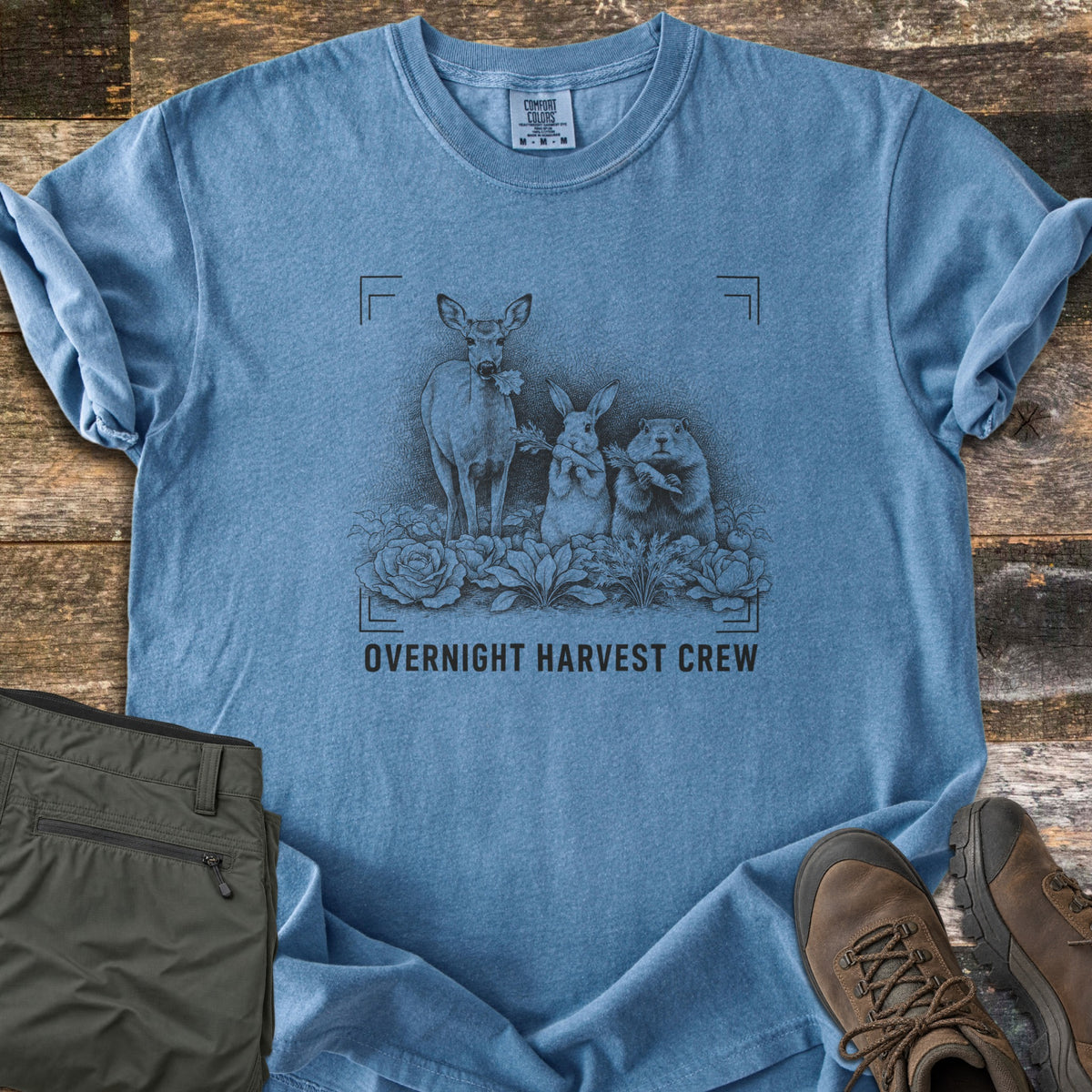 Overnight Harvest Crew Comfort Colors T-shirt in Blue Jean, laid flat. A dark sketch of a deer chewing a leaf, a rabbit and a groundhog holding carrots in a cabbage patch, inside camera frame corners above the words Overnight Harvest Crew. Olive pants and brown boots lie on wood.